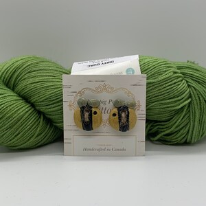 May include: A pair of handmade ceramic bee earrings with black stripes, yellow bodies, and green accents. The earrings are displayed on a white card with the text "The Big Potto" and "Handcrafted in Canada".