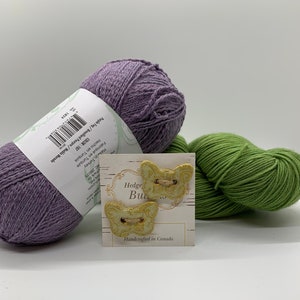 Puede incluir: Dos botones en forma de mariposa de color amarillo y verde con detalles dorados en una tarjeta blanca con el texto "Hedge But" y "Handcrafted in Canada". La tarjeta está encima de dos ovillos de lana, uno morado y otro verde.
