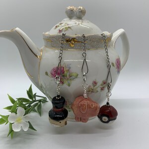 May include: Three decorative tea infusers suspended from a white teapot. The infusers feature a black and red top hat, a pink pig, and a red and white ball. The teapot has a floral design and gold trim.