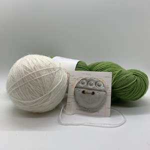 Puede incluir: Un botón de cerámica gris con un diseño texturizado, sujeto a una etiqueta con el texto "Handcrafted in Canada". Una bola de hilo blanco y un ovillo de hilo verde también están en la imagen.