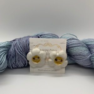 May include: Two white ceramic flower-shaped buttons with gold accents. The buttons are on a white card with the text "Hedgepig Pottery, Ltd. Handcrafted in Canada".