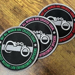 May include: Three round black patches with white and colored text. Each patch features a pair of handcuffs and the text "NOT ALL WOMEN ARE CREATED EQUAL" and "THE BRAVEST BECOME LAW ENFORCEMENT OFFICERS". The text is in white, green, and pink.