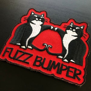 May include: Red embroidered patch with two black and white cats with white faces and black eyes. The cats are holding tails and the text "Fuzz Bumper" is embroidered in white on the patch.