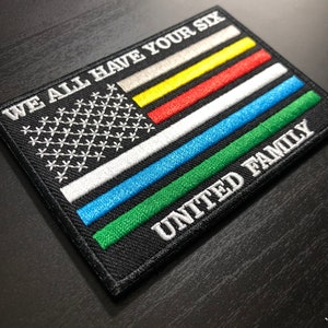 May include: Embroidered patch featuring a stylized American flag design with the text "WE ALL HAVE YOUR SIX" and "UNITED FAMILY." The flag incorporates various colored stripes, including gray, yellow, red, blue, and green, on a black background.