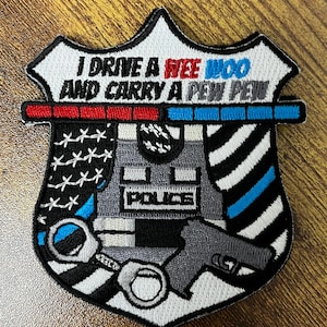 May include: Embroidered police badge patch with the text "I DRIVE A WEE WOO AND CARRY A PEW PEW." The patch features a shield design with a police vest, handcuffs, and a gun. The patch has a white border and is on a wooden surface.