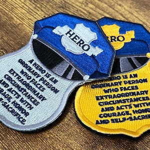May include: Two embroidered patches in the shape of police badges. One is blue and silver, the other yellow and black. Both have the word "HERO" and a quote about heroes. The patches are on a wooden surface.