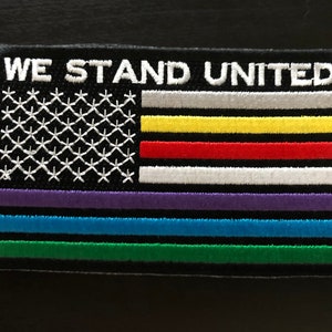 May include: An embroidered patch with the words "WE STAND UNITED" above a stylized American flag with multiple colored stripes. The stripes are purple, blue, green, white, red, yellow, and gray.