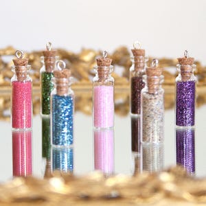 May include: A collection of small glass bottles filled with colorful glitter. Each bottle has a cork stopper and a silver loop at the top. The glitter colors include pink, green, blue, pink, brown, beige, and purple. The bottles are displayed on a reflective surface.