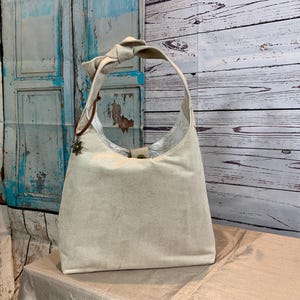 May include: A beige canvas tote bag with a knotted handle and a decorative metal embellishment. The bag is sitting on a light brown surface.