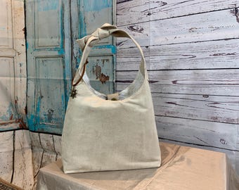 May include: A beige canvas tote bag with a knotted handle and a decorative metal embellishment. The bag is sitting on a light brown surface.