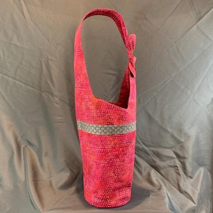 Pode incluir: Uma bolsa alta de tecido rosa com uma única alça de ombro. A bolsa tem um padrão texturizado e uma faixa decorativa de tecido cinza e branco. A alça é amarrada no topo. A bolsa tem aproximadamente 51 cm de altura.