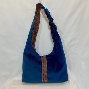 Pode incluir: Uma bolsa hobo azul profundo com uma alça de ombro única com detalhe de nó. O exterior é de tecido azul sólido, enquanto o forro interno apresenta um padrão floral marrom. Uma faixa vertical de acabamento floral acentua o centro da frente.
