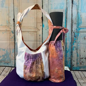 May include: A white canvas yoga bag with a purple and orange floral print pocket. The bag has a long strap and is holding a rolled up black yoga mat.