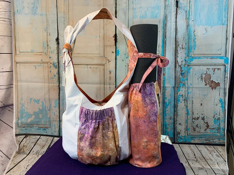 May include: A white canvas yoga bag with a purple and orange floral print pocket. The bag has a long strap and is holding a rolled up black yoga mat.