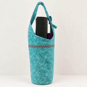 Pode incluir: Uma bolsa de tecido verde-azulado para tapete de ioga com um padrão floral em espiral e uma alça de ombro atada no topo. Uma faixa horizontal cinzenta com um design geométrico repetitivo envolve a bolsa que contém um tapete preto.