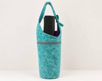 May include: A teal fabric yoga mat bag with a swirling floral pattern and a single shoulder strap tied at the top. A gray horizontal band with a repeating geometric design encircles the bag, which contains a black yoga mat.