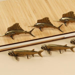 May include: Bronze-colored fish-shaped decorative hardware. The image shows six fish, three with tall dorsal fins and three with shorter fins. They are arranged on a wooden surface.