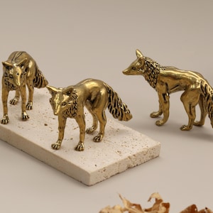 May include: Three brass fox figurines. Two are on a rectangular stone, and one is separate. The foxes have detailed fur and are in different poses. The background is a neutral colour.