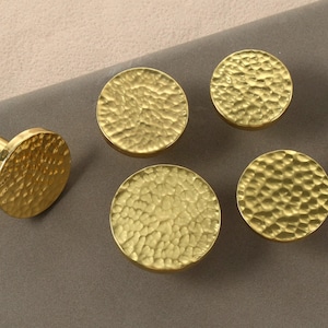 Puede incluir: Un conjunto de cinco tiradores dorados para armarios con textura martillada. Los tiradores son redondos, con un tirador que tiene una base cilíndrica. Los tiradores se muestran sobre una superficie gris.