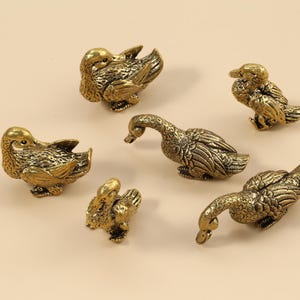 May include: A selection of small, detailed brass duck figurines in various poses. The ducks are in different positions, some with their heads down, others upright. The brass has a warm, antique finish, and the ducks are arranged on a neutral background.