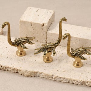 May include: Three brass-coloured dinosaur figurines with long necks and flipper-like appendages are displayed on a textured, light-coloured surface. The figurines are mounted on round bases and are positioned in front of off-white, geometric blocks.
