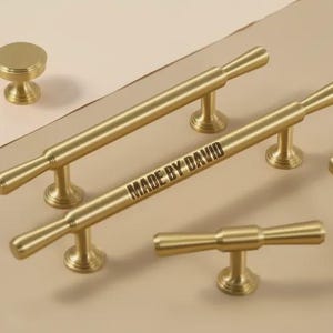 May include: Gold-coloured cabinet hardware set, including a knob, bar pulls and a T-pull. The bar pulls have the text "MADE BY DAVID" engraved on them. The hardware is displayed on a neutral surface.