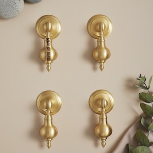 Puede incluir: Cuatro tiradores de armario de color latón con una placa trasera redonda y un asa en forma de lágrima. Un tirador tiene el texto "MADE BY DAVID" en el asa. Los tiradores están montados en una pared beige, con piedras grises y vegetación.