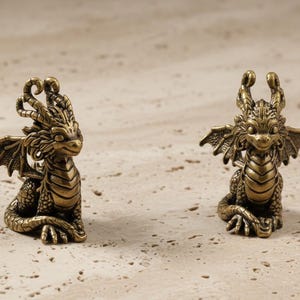 May include: Two bronze dragon figurines. Each dragon has detailed wings, a spiky spine, and a friendly expression. The dragons are posed sitting upright, with their wings slightly spread. The background is a light, textured surface.