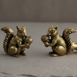 May include: Two antique brass squirrel figurines, each holding a nut. The squirrels have detailed fur and tail textures. The figurines are on a grey surface, with a neutral background. The squirrels are a decorative item.