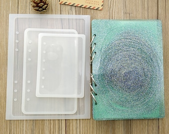 Può includere: Tre stampi in silicone trasparente per realizzare un quaderno a spirale. Gli stampi sono annidati uno dentro l'altro, con lo stampo più grande in basso e quello più piccolo in alto. Gli stampi sono su una superficie di legno. Accanto agli stampi c'è un quaderno a spirale con glitter turchese.
