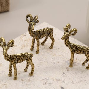 Puede incluir: Tres pequeñas figuras de carnero de color oro antiguo. Cada carnero tiene cuernos detallados y se apoya sobre cuatro patas. Las figuras se exhiben sobre una superficie clara y texturizada, lo que sugiere un tema decorativo o de colección.