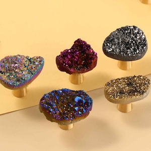 May include: Five cabinet knobs featuring irregular, geode-style tops with crystalline textures in iridescent, deep purple, dark gray, blue, and gold tones. Each knob is mounted on a cylindrical, brushed gold-colored metal base for attachment.