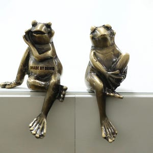 Puede incluir: Dos figuras de rana de bronce, cada una posada sobre una superficie blanca. Una rana apoya la barbilla en la mano, mientras que la otra está sentada con los brazos cruzados. El texto "MADE BY DAVID" es visible en una rana. Esculturas decorativas.