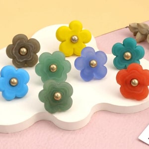May include: A collection of eight flower-shaped knobs in various colors including yellow, blue, green, orange, and brown. Each knob features a five-petal design with a central metallic gold-toned circular accent, providing a uniform look.