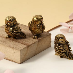 以下が含まれることがあります： 3つの小さな真鍮製のフクロウの置物。2羽のフクロウが小さな正方形の木製ブロックに止まっています。フクロウは大きな目と質感のある羽を持っています。背景は、ピンクの花がある柔らかいニュートラルカラーです。