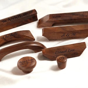 May include: A collection of wooden cabinet hardware, including handles and knobs. Each piece is a rich brown color and engraved with names or phrases like "Annie," "Charlotte," and "Eternal Love." The hardware is displayed on a white surface.