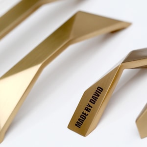 May include: Gold-toned cabinet pulls with a geometric design. The pulls have a faceted shape and a smooth, metallic finish. One pull is labeled "MADE BY DAVID" in black text. These are likely for furniture or cabinetry.