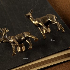 May include: Two bronze-colored animal-shaped cabinet knobs. One is a moose, the other a deer. The knobs are mounted on a dark surface. The moose knob has antlers, and the deer knob has a slender neck.