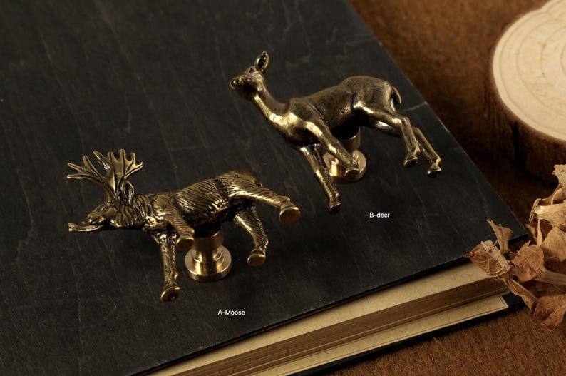 May include: Two bronze-colored animal-shaped cabinet knobs. One is a moose, the other a deer. The knobs are mounted on a dark surface. The moose knob has antlers, and the deer knob has a slender neck.
