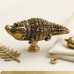 May include: A brass cabinet knob shaped like a crocodile with detailed scales, accompanied by a brass fern leaf, two brass acorn-shaped knobs, and a small hexagonal nut. A portion of a metal ruler is visible at the bottom of the frame.