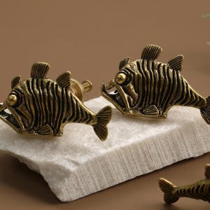 May include: Two antique brass fish-shaped cabinet knobs with open mouths and detailed fins rest on a white stone. The knobs are a decorative hardware element, adding a unique touch to furniture.