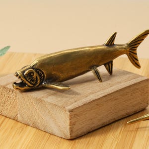 Op de afbeelding: Een bronzen visfiguur met een open mond, geplaatst op een klein houten blok. De vis heeft gedetailleerde vinnen en een getextureerd lichaam. De figuur staat op een houten oppervlak, geschikt voor een nautisch of decoratief thema.