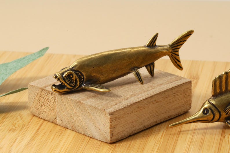 Op de afbeelding: Een bronzen visfiguur met een open mond, geplaatst op een klein houten blok. De vis heeft gedetailleerde vinnen en een getextureerd lichaam. De figuur staat op een houten oppervlak, geschikt voor een nautisch of decoratief thema.
