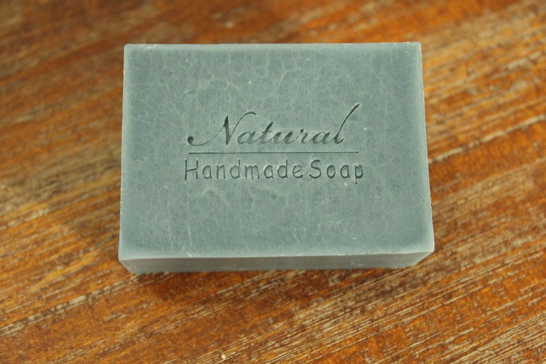 Op de afbeelding: Een rechthoekige blok handgemaakte zeep in een gedempte blauwgroene kleur. De woorden "Natural Handmade Soap" zijn op het oppervlak ge&euml;mbosseerd. De zeep wordt gepresenteerd op een houten oppervlak, wat de natuurlijke uitstraling benadrukt.