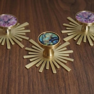 Può includere: Tre pomelli circolari per mobili con una piastra posteriore in metallo dorato spazzolato a forma di raggiera. Due pomelli presentano un intarsio in pietra marmorizzata viola e bianca, mentre quello centrale ha un intarsio in conchiglia abalone.