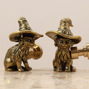 May include: Two brass cat figurines wearing witch hats. The cats are detailed with textured fur and are posed in a sitting position. The hats have a classic witch hat shape with a wide brim. The figurines are likely decorative items.