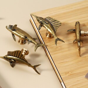 Op de afbeelding: Vier antieke messing visvormige kastknoppen. De knoppen zijn gedetailleerd met vinnen, staarten en een lange, puntige snuit. Ze zijn gemonteerd op een lichtgekleurd houten oppervlak, met een neutrale achtergrond. Decoratief en functioneel.