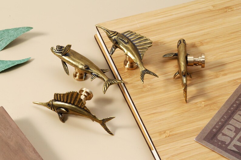 Op de afbeelding: Vier antieke messing visvormige kastknoppen. De knoppen zijn gedetailleerd met vinnen, staarten en een lange, puntige snuit. Ze zijn gemonteerd op een lichtgekleurd houten oppervlak, met een neutrale achtergrond. Decoratief en functioneel.