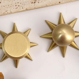 以下が含まれることがあります： 8つの尖った光線を持つスターバーストデザインのゴールドカラーの金属製キャビネットノブのセット。1つのノブは同心円状のリングがある平らな円形の面を持ち、もう1つは滑らかで丸みを帯びたドーム状の中心部を持っています。