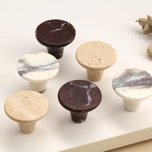 Puede incluir: Un conjunto de seis pomos circulares para armarios con texturas de piedra variadas. El set incluye dos pomos de color marrón oscuro con vetas blancas, dos pomos de piedra beige porosa, un pomo blanco con vetas grises y moradas, y uno blanco con vetas verdes.